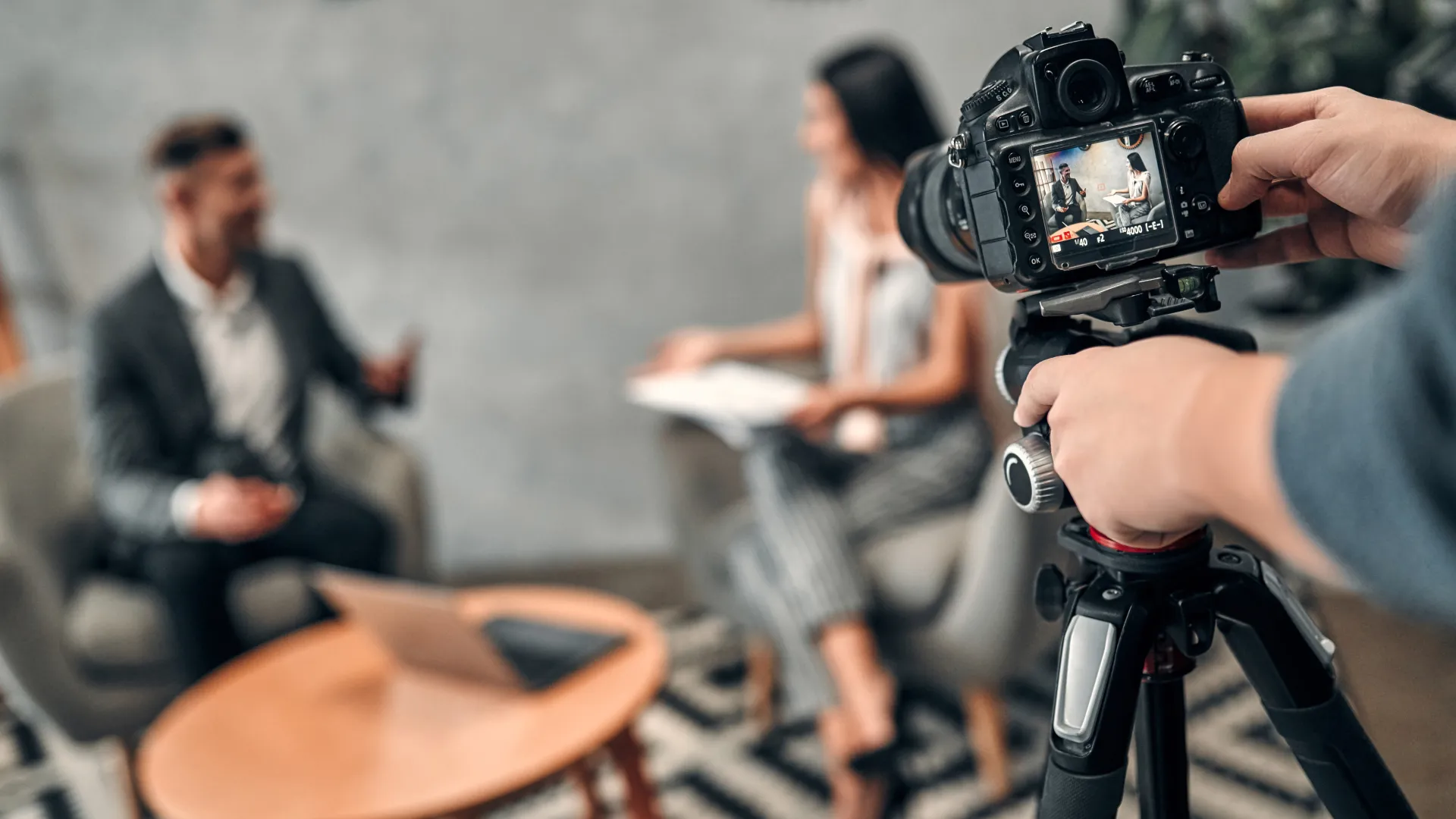 Im Vordergrund ein Stativ mit Kamera, im Hintergrund findet ein Interview statt, unscharf zu sehen sind eine weibliche und eine männliche Person in einer Interviewsituation.